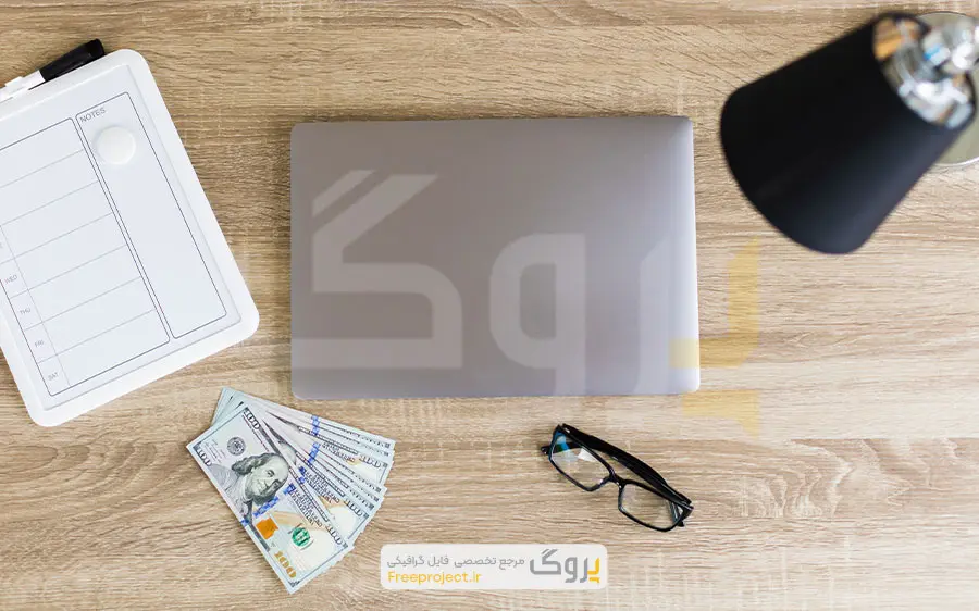 عکس استوک نمای بالا از میز کار با لپ‌تاپ، عینک، چراغ و اسکناس‌های دلار
